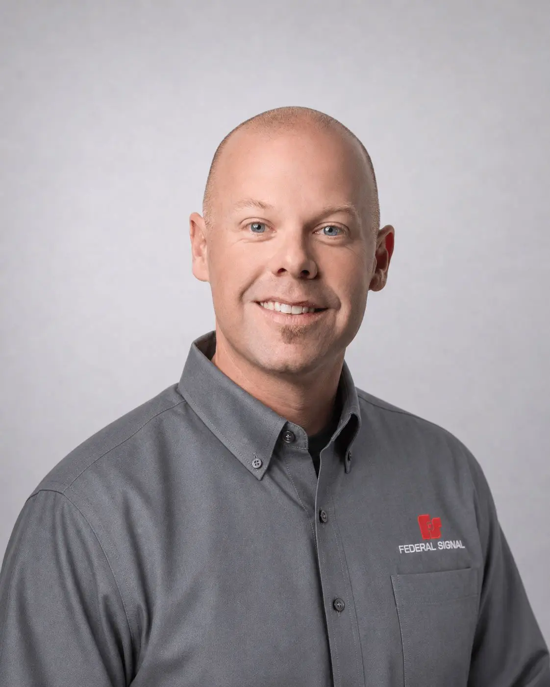 A man with a shaved head wearing a gray Federal Signal branded shirt poses and smiles in front of a plain light gray background.