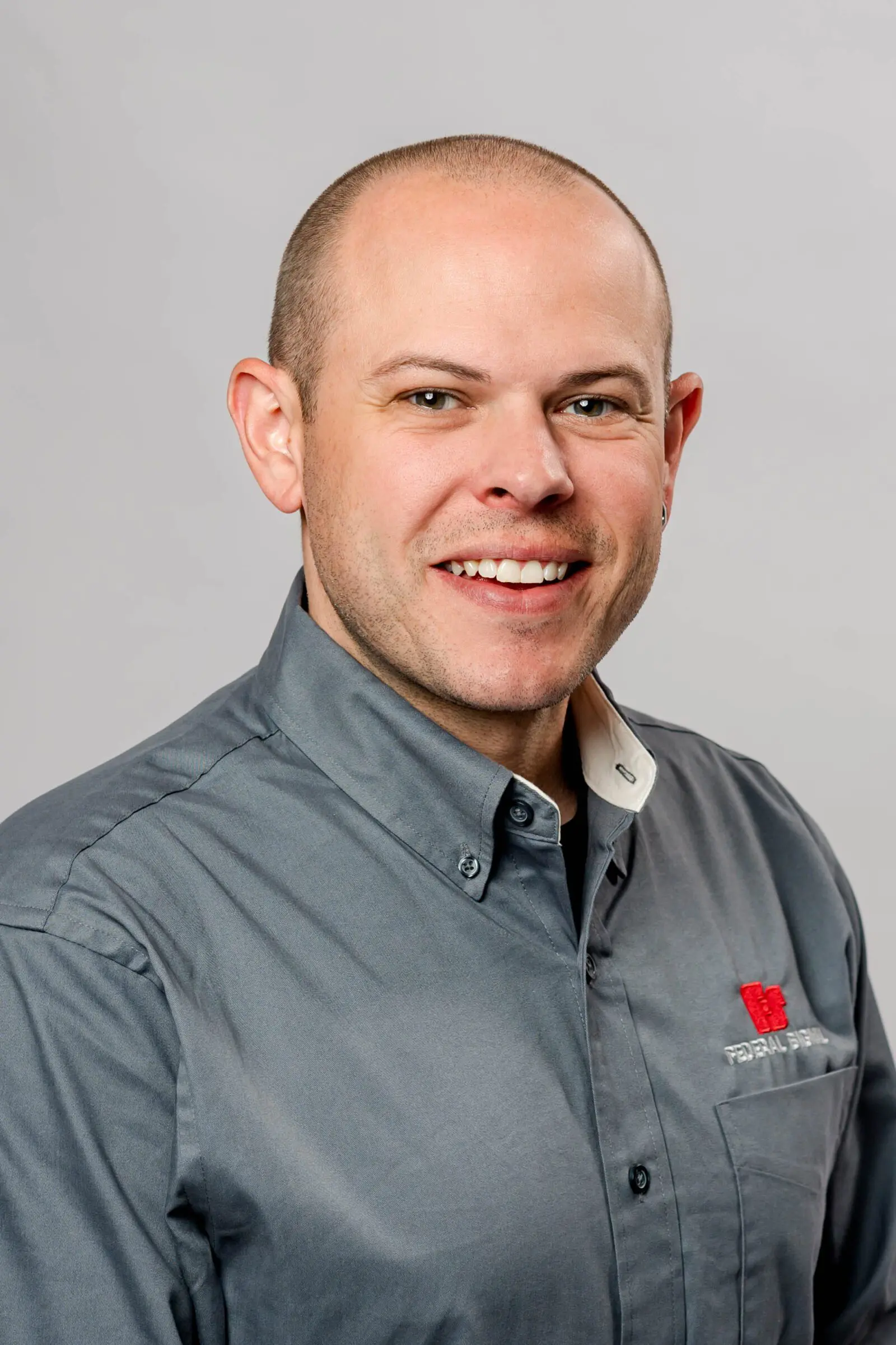 A smiling man with a shaved head wearing a gray button-up shirt featuring a red and white embroidered logo.