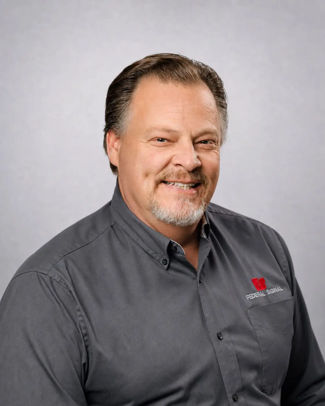 A middle-aged man with short hair and a goatee, wearing a gray collared shirt with a “Federal Signal” logo, poses against a plain light background.