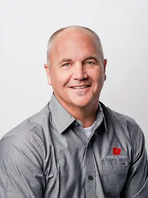 A man with short gray hair is smiling, wearing a gray collared shirt with a logo on the left side.
