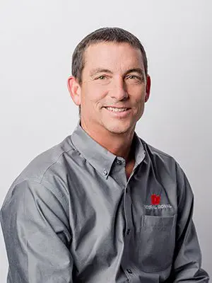 A man in a gray shirt with a red logo sits smiling in front of a plain background.