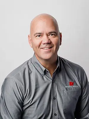 A bald man wearing a gray shirt with a red logo smiles at the camera against a plain background.