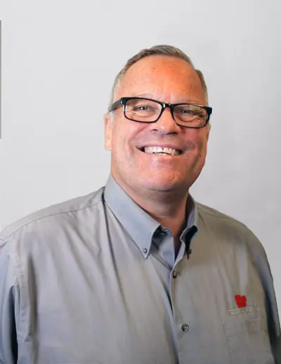 Man with glasses smiling, wearing a gray button-up shirt with a red logo. Neutral background.