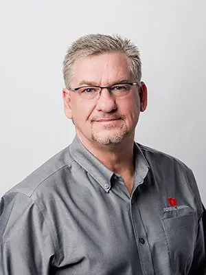 Man with glasses and short gray hair wearing a gray button-up shirt with a logo on the chest, standing against a plain gray background.