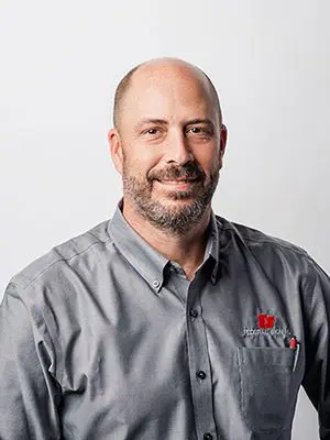 A person with a beard and mustache, wearing a gray collared shirt with a logo, smiles at the camera against a plain background.