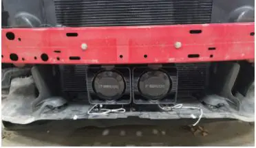 Close-up of a car's front bumper showing two fans secured to the radiator support behind a red metal bar.