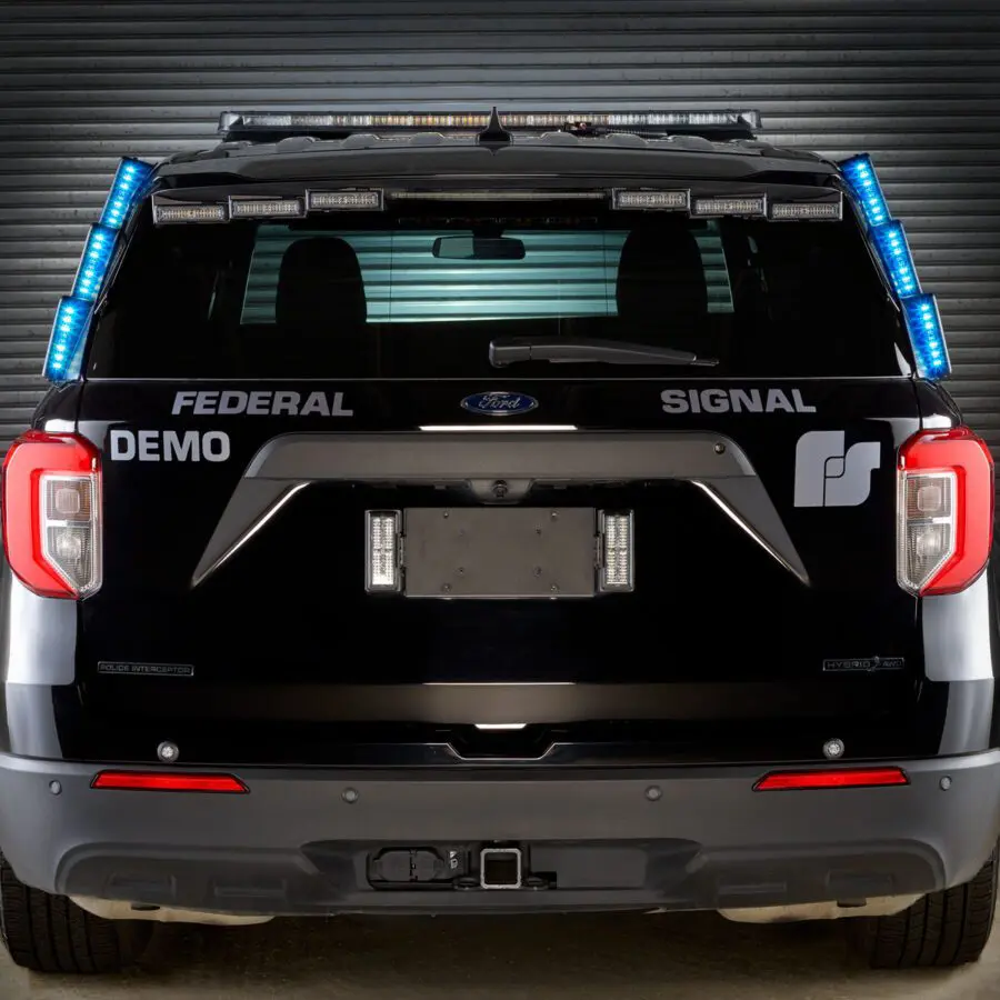 A rear view of a black SUV equipped with FedSig's MicroPulse Ultra D-Pillar, featuring blue and white emergency lights activated, is parked in front of a closed garage door.