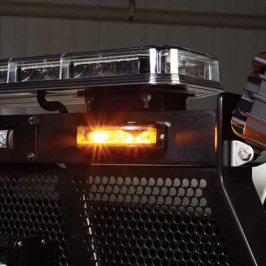 Close-up of a FedSig MicroPulse Wide Angle amber emergency light bar illuminated against a black metal grille background.