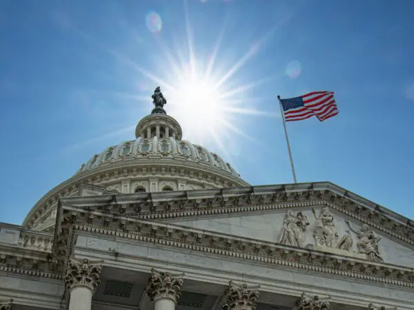 The U.S. Capitol building dome with a bright sunburst overhead and an American flag flying on the right.