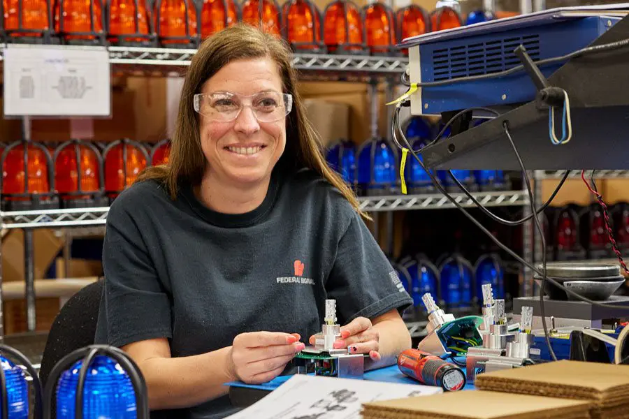 Woman wearing safety glasses assembling electrical components in a workshop, surrounded by red and blue lights on shelves.