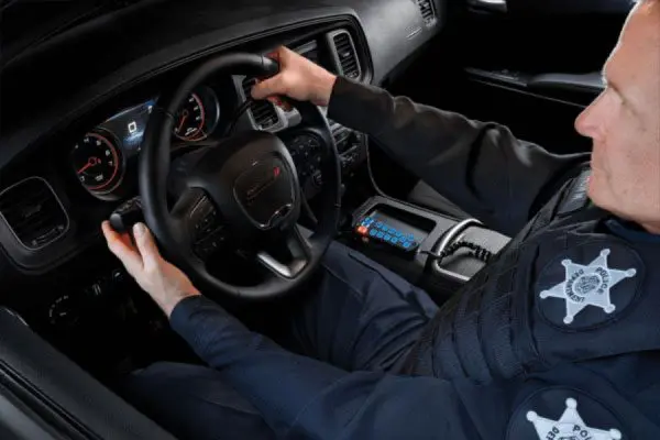 A police officer is in a patrol car, holding the steering wheel with one hand while adjusting a control on the dashboard with the other.