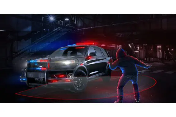 A person in a hoodie stands in front of a police SUV with flashing lights at night, highlighting its surveillance technology.