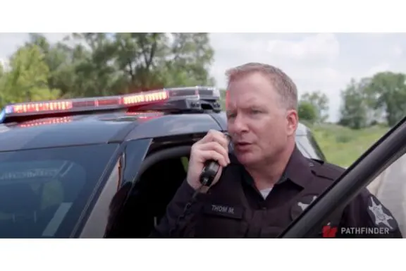 Police officer speaking into a radio, standing next to a patrol car with lights on. Trees and cloudy sky in the background.