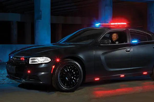 A black police car with illuminated red and blue lights is parked under a bridge at night.