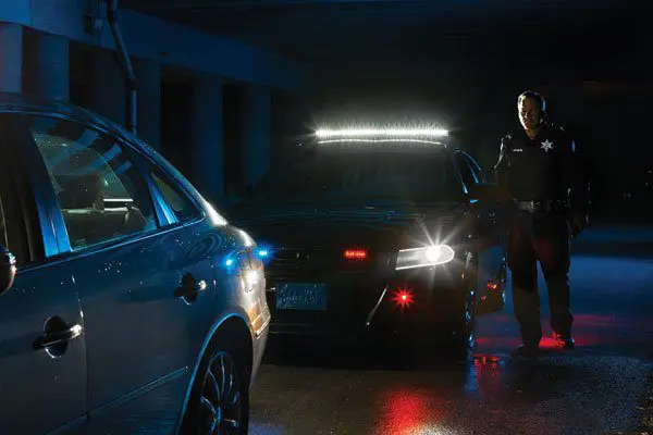 Police officer stands next to a patrol car with flashing lights behind a stopped vehicle in a dimly lit area.