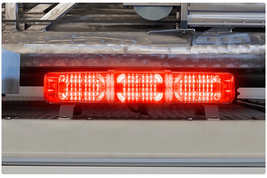 Red emergency vehicle light bar illuminated, mounted on a metallic surface with industrial equipment in the background.