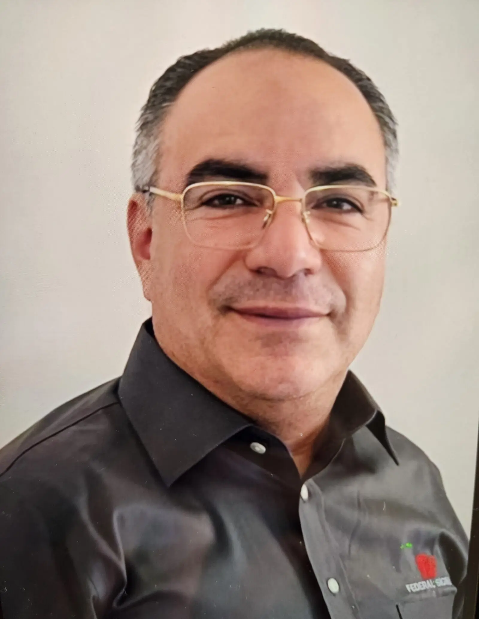 A middle-aged man with short gray hair, glasses, and a dark button-up shirt, facing the camera and smiling slightly against a plain background.