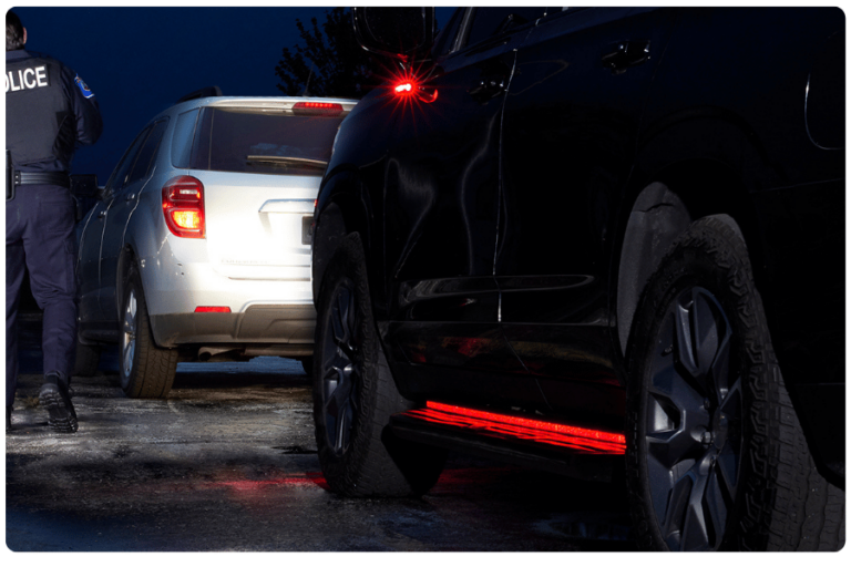 A police officer stands beside two cars stopped on a wet road at night, with the vehicles' brake lights illuminated.