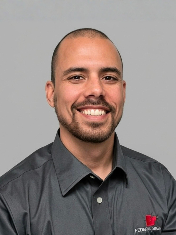 A man with a shaved head and short beard smiles at the camera, wearing a dark button-up shirt with an embroidered red and white logo on the chest.