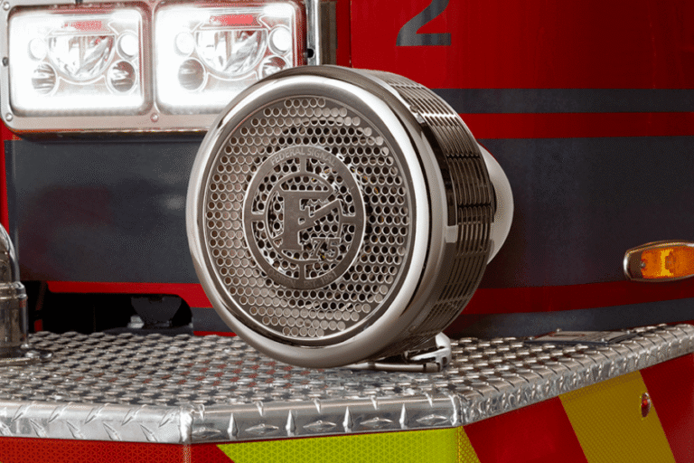 A close-up of a chrome fire truck siren mounted on a metal diamond plate surface, with part of the red fire truck and emergency lights visible in the background.