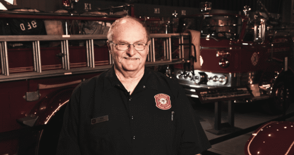 An older man wearing a fire department shirt stands in front of a vintage fire truck inside a fire station.
