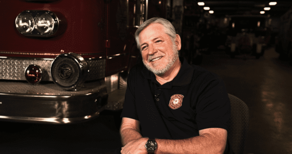An older man with gray hair and beard, wearing a fire department polo shirt, sits and smiles in front of a fire truck inside a fire station.