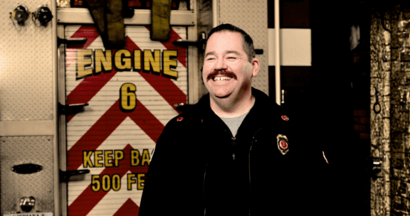 A smiling firefighter stands in front of a fire truck with “Engine 6” and “Keep Back 500 Feet” written on the back.