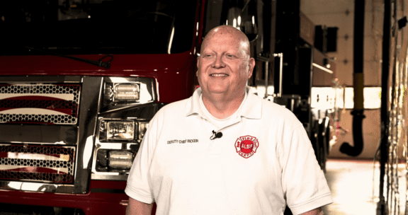 A man in a white "Deputy Chief Richer" shirt stands smiling in front of a fire truck inside a fire station.