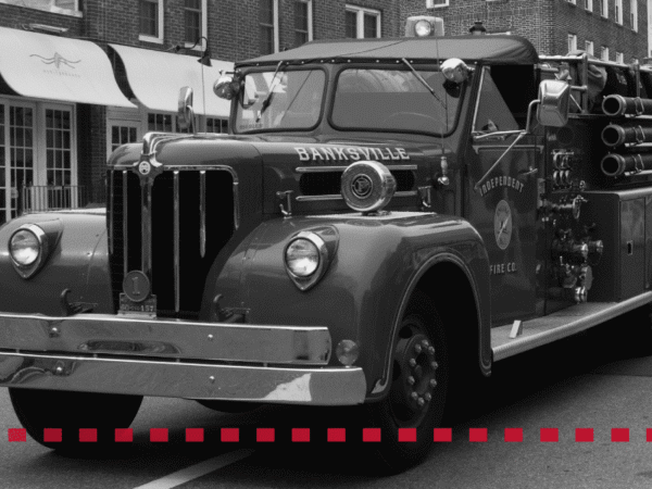 A vintage Banksville fire truck is parked on a street in front of storefronts. The image is in black and white with a dashed red line across the bottom.