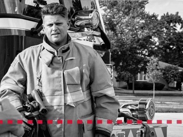 A firefighter in uniform stands in front of a fire truck, holding equipment, with houses and trees visible in the background.
