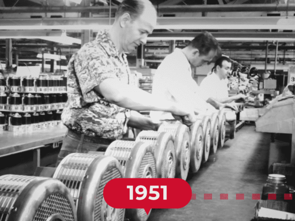 Three men work on an assembly line in a factory, handling circular metal components; a "1951" label is displayed in the foreground.