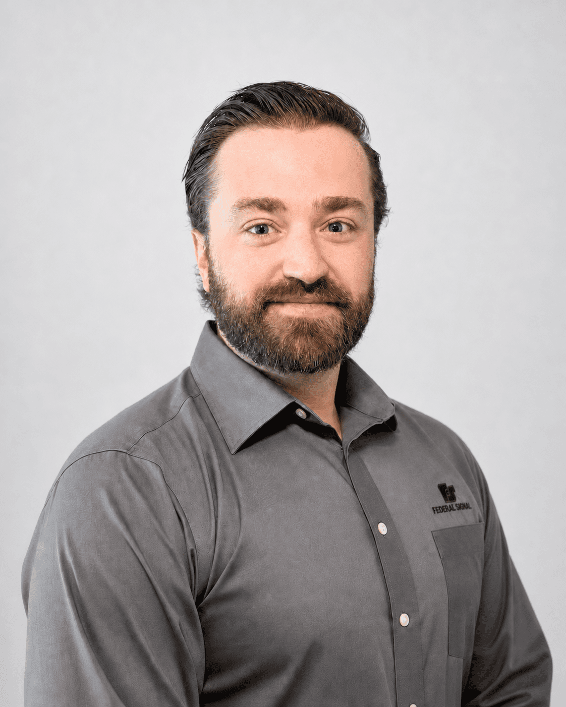 A man with dark hair and a beard, wearing a gray collared shirt with a logo, stands against a plain light gray background, facing the camera and smiling slightly.