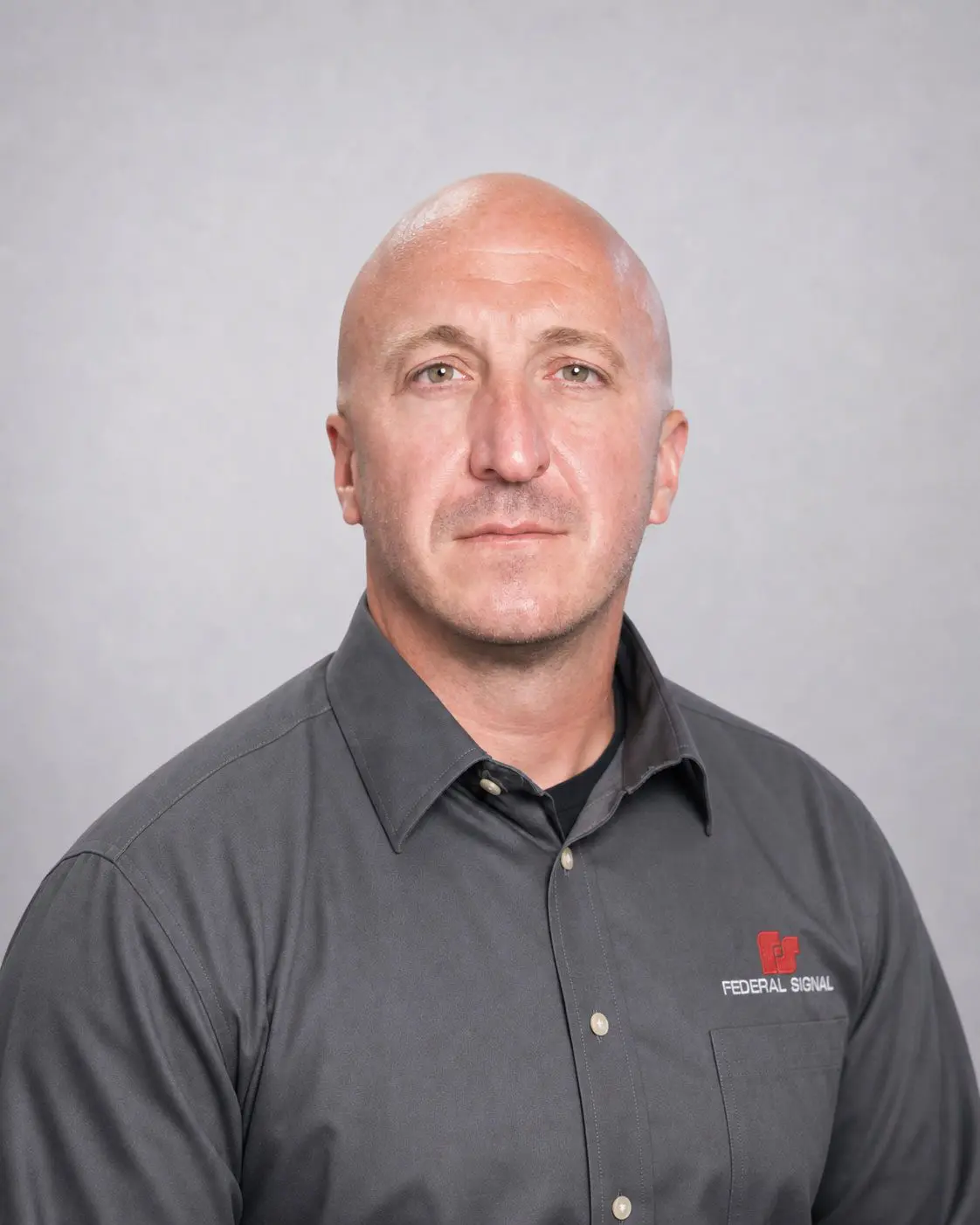 A man with a shaved head wearing a gray “Federal Signal” shirt poses for a professional portrait against a plain light gray background.