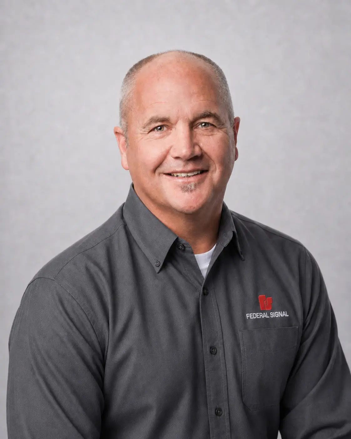 Smiling middle-aged man in a gray “Federal Signal” shirt, sitting in front of a plain light gray background.