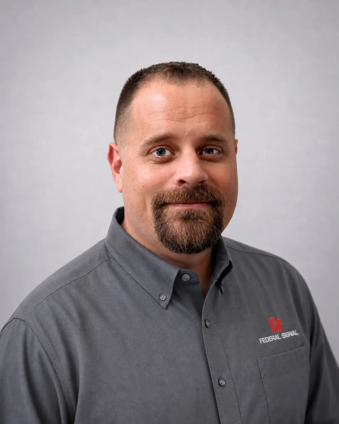A man with short hair and a goatee wearing a grey collared shirt with a Federal Signal logo, facing the camera against a plain light background.