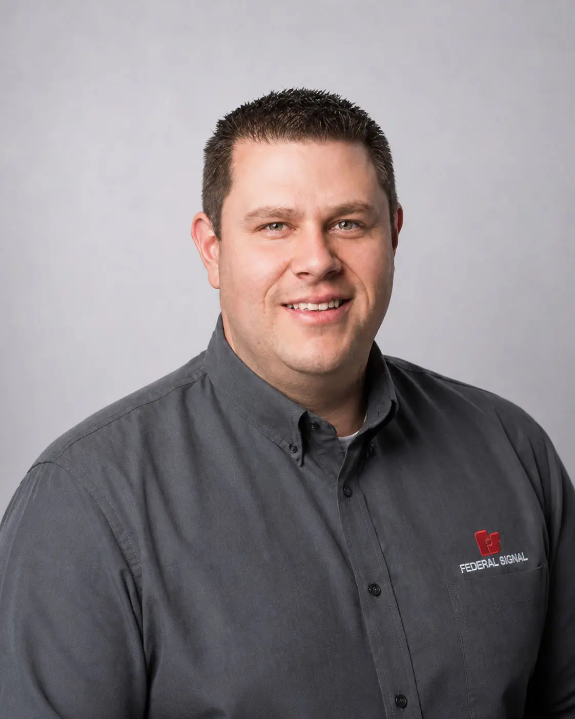 Man with short brown hair wearing a gray “Federal Signal” button-down shirt, smiling, standing against a plain light gray background.