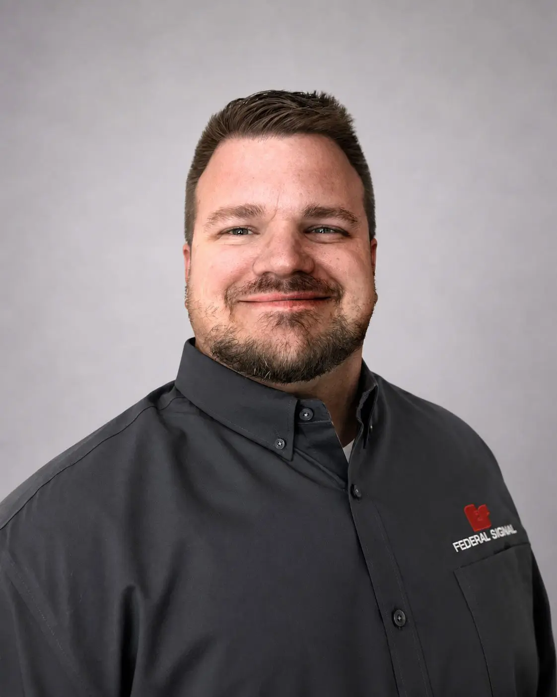 A man with short brown hair and a beard smiles, wearing a dark gray collared shirt with the Federal Signal logo, against a plain light gray background.