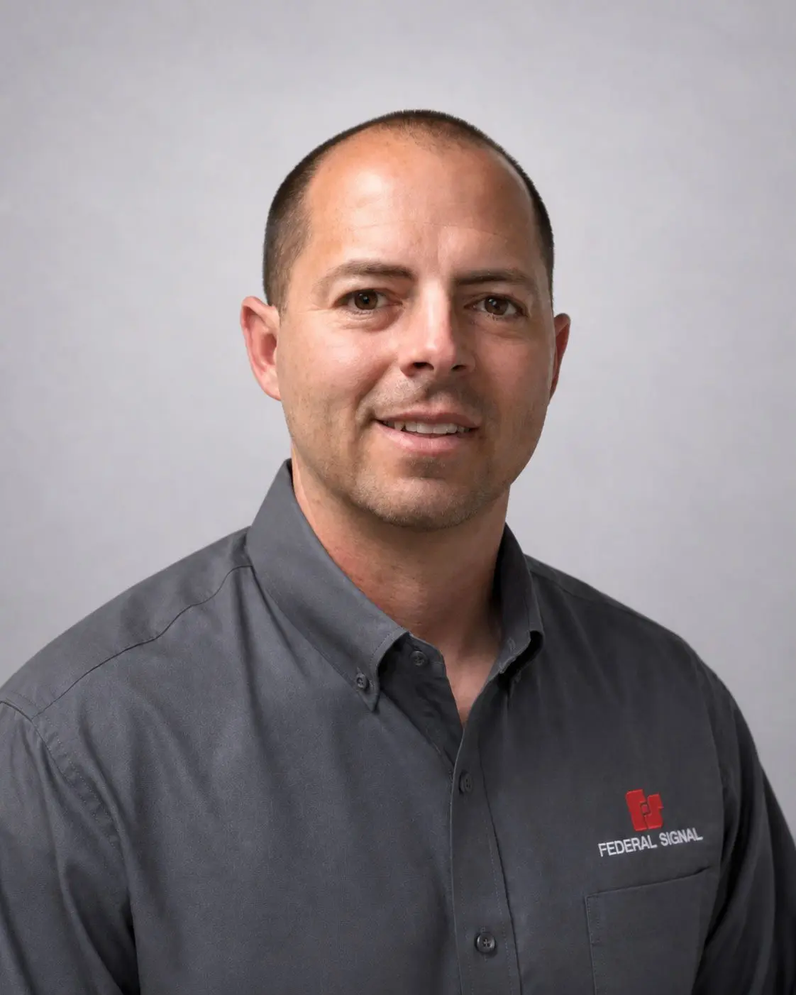 A man with short hair wearing a gray “Federal Signal” button-up shirt poses for a professional headshot against a light gray background.