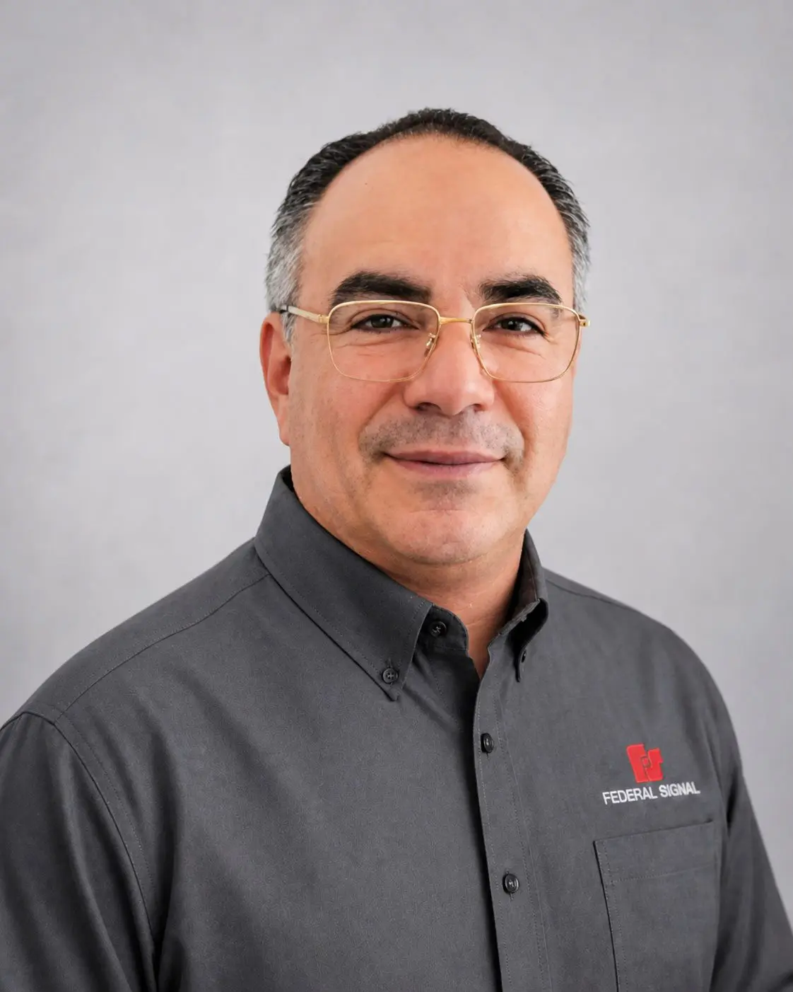Man wearing glasses and a dark gray shirt with a Federal Signal logo, standing in front of a plain light gray background.