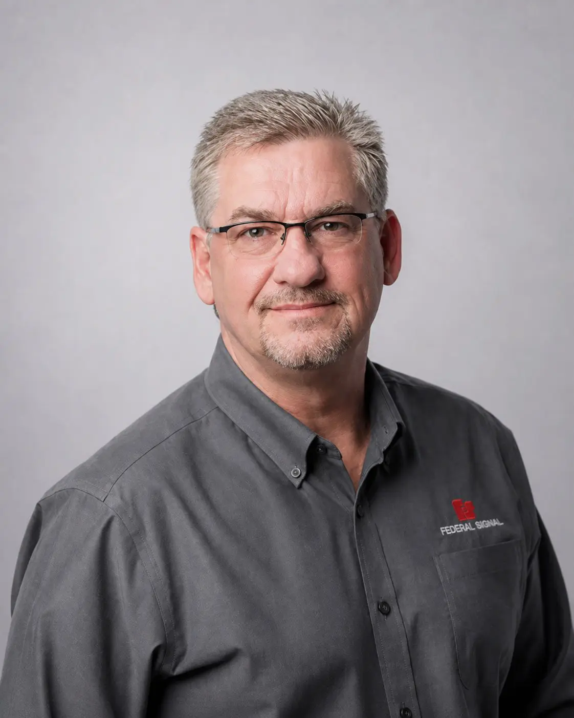 Middle-aged man with short gray hair, glasses, and a goatee wearing a gray button-up shirt with a “Federal Signal” logo, posing against a plain light gray background.