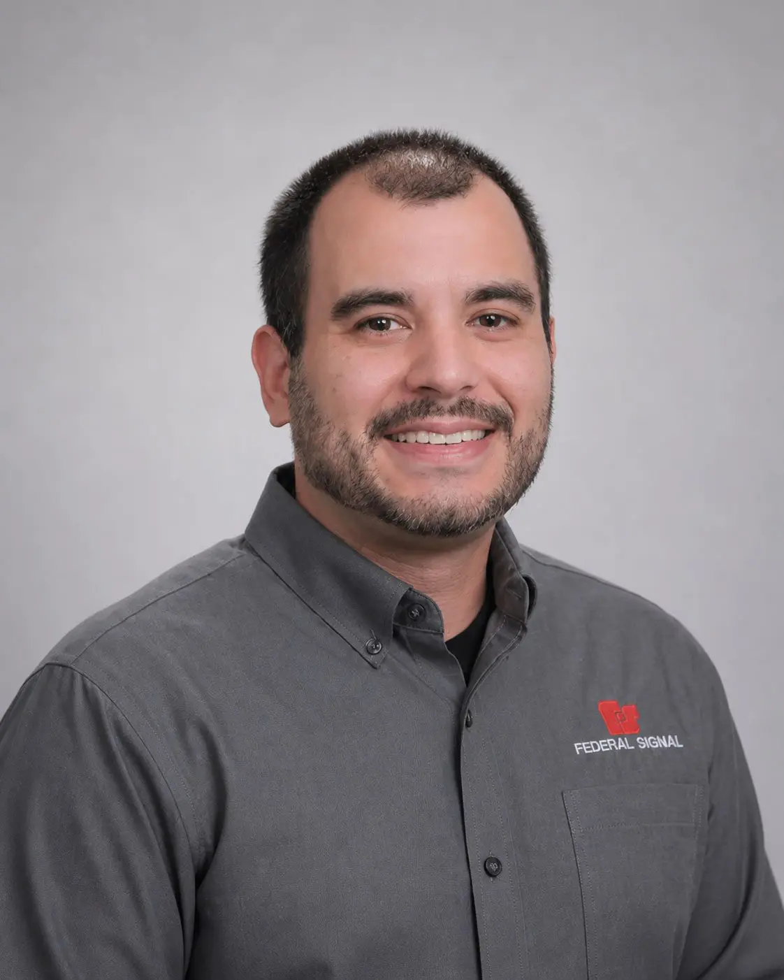 Man with short dark hair and beard smiling, wearing a gray collared shirt with a red “Federal Signal” logo on the left chest, against a plain light gray background.